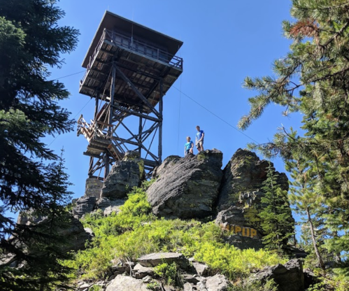 A tall lookout tower atop rocky terrain, surrounded by trees and greenery, with two figures standing on the rocks.