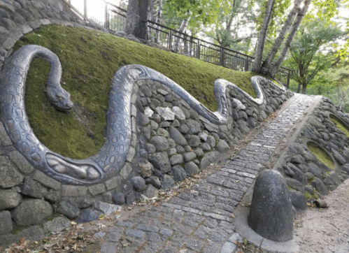 A winding, metallic snake sculpture on a moss-covered stone wall beside a cobblestone path in a wooded area.