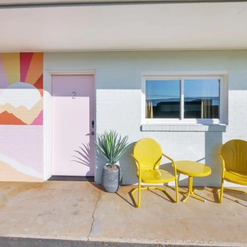 Colorful exterior of a building featuring a pink door, potted plant, and yellow chairs on a sunny day.