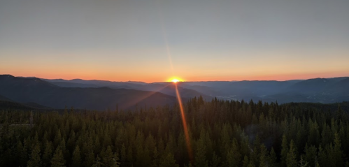 A stunning sunset over a mountain range, with vibrant orange and pink hues illuminating the sky above a forested landscape.