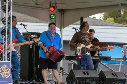 A band performs on stage under a tent, featuring a guitarist, accordion player, and bassist with musical equipment.