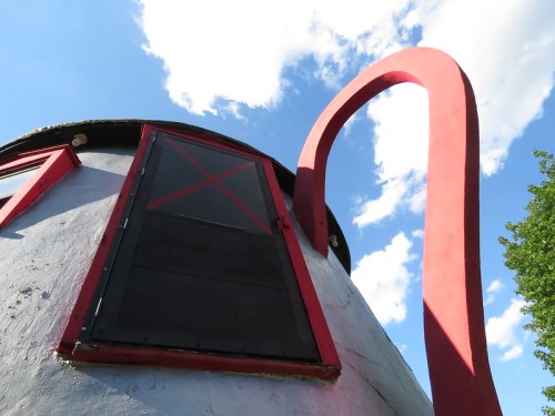 A close-up view of a whimsical building with a curved red structure and a black window against a blue sky.