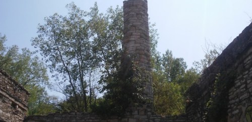 Ruins of a stone structure with a tall, cylindrical chimney surrounded by trees under a clear blue sky.