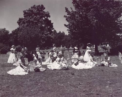 A group of women in vintage dresses poses in a cemetery, surrounded by gravestones and trees on a sunny day.