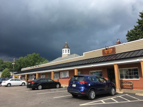 A pizza restaurant in a shopping plaza under a dark, cloudy sky, with parked cars in the foreground.