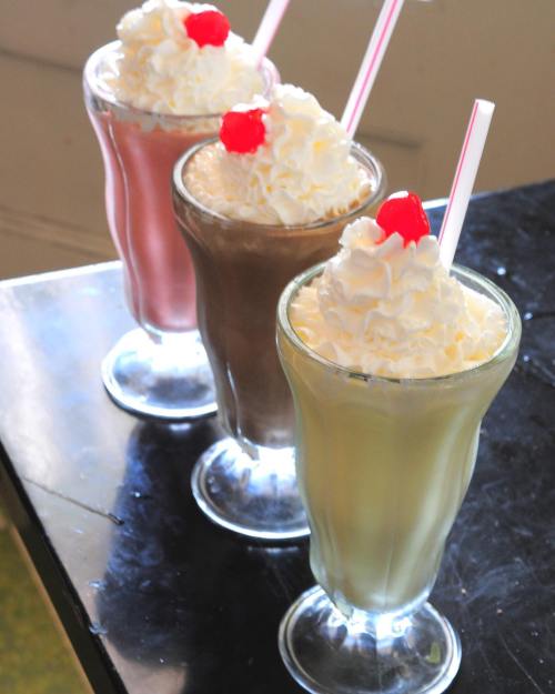 Three milkshakes in tall glasses, topped with whipped cream and cherries, in pink, brown, and green colors.
