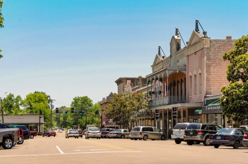 A sunny street scene featuring historic buildings, parked cars, and trees lining the road in a small town.