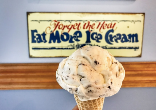 A hand holding a cone of ice cream with a sign in the background that says, "Forget the Year, Eat More Ice Cream."