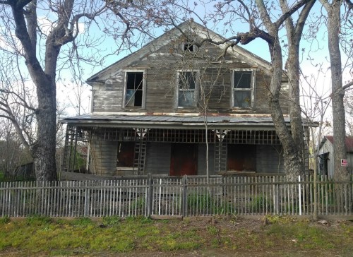Abandoned two-story house with peeling paint, broken windows, and a wooden fence, surrounded by bare trees.