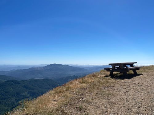 A picnic table on a grassy hilltop overlooking a vast mountain landscape under a clear blue sky.
