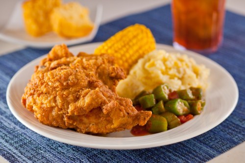 A plate of fried chicken with corn on the cob, mashed potatoes, and okra, served on a blue placemat.