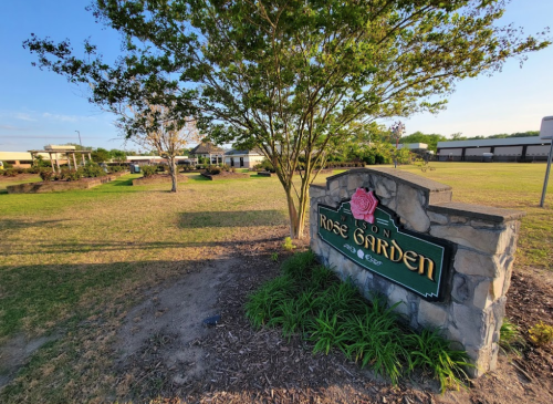 A stone sign reading "Rose Garden" in a landscaped area with trees and buildings in the background under a clear sky.