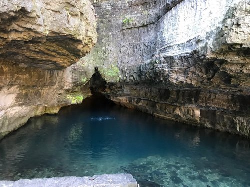 A serene cave entrance with dark rock walls and clear blue water reflecting the light.