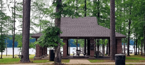 A pavilion surrounded by trees near a lake, with picnic tables and trash bins visible.