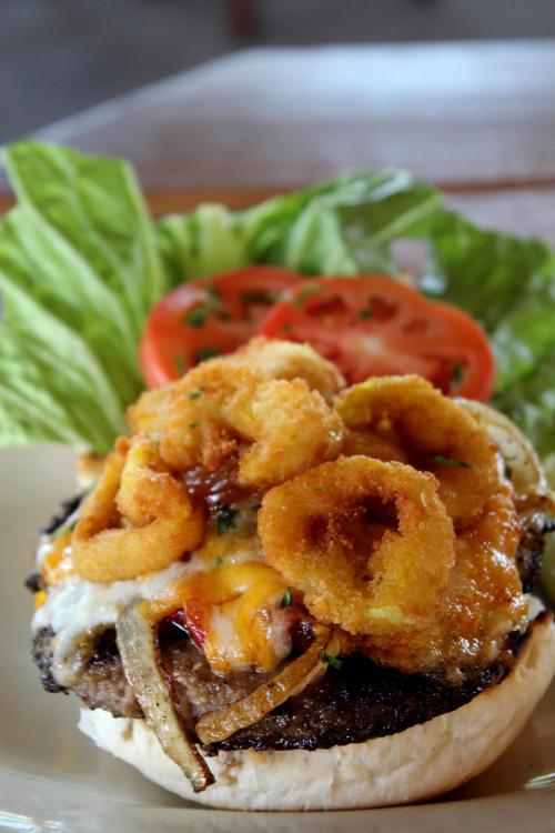 A close-up of a gourmet burger topped with cheese, fried onion rings, lettuce, and tomato on a toasted bun.