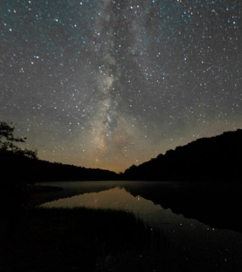 A serene night sky filled with stars and the Milky Way reflected in a calm lake, surrounded by dark silhouettes of trees.