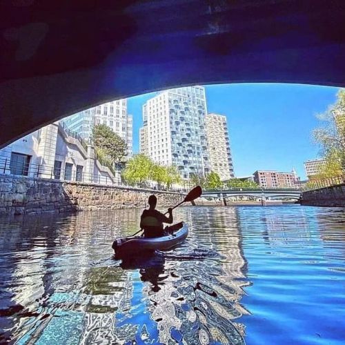 A person kayaking under a bridge, surrounded by city buildings and reflecting water on a sunny day.