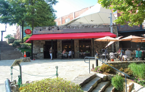 Outdoor view of Passerelle Bistro with a red awning, surrounded by greenery and patrons dining at tables.