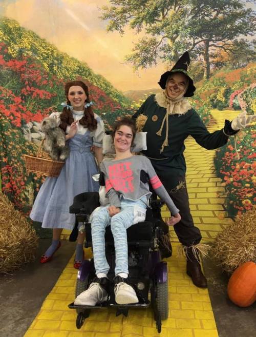 A girl in a wheelchair smiles with Dorothy and the Scarecrow on a yellow brick road backdrop.