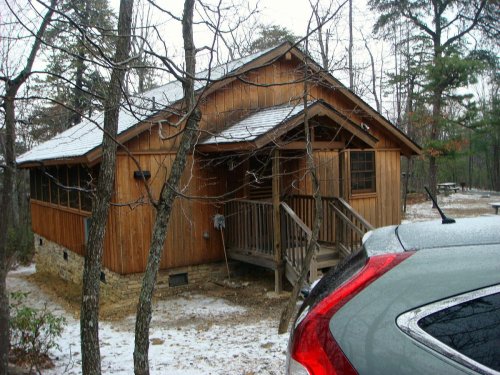 A wooden cabin surrounded by trees, with a sloped roof and a small porch, partially covered in snow.