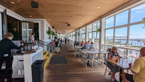 A bright restaurant interior with wooden floors, large windows, and diners enjoying meals by the water.