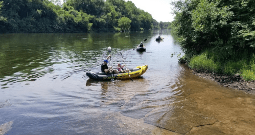 Two people in a yellow raft on a calm river, with others in kayaks nearby and lush greenery along the shore.