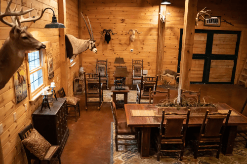 A rustic dining room with wooden walls, a large table, chairs, and mounted animal heads on the walls.