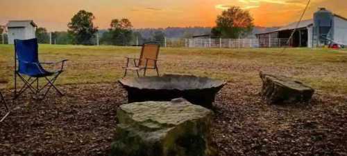 A serene outdoor scene with a fire pit, two chairs, and a sunset over a grassy field and barn in the background.