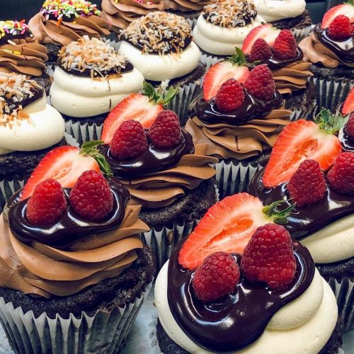 A close-up of assorted cupcakes topped with chocolate frosting, strawberries, raspberries, and coconut flakes.