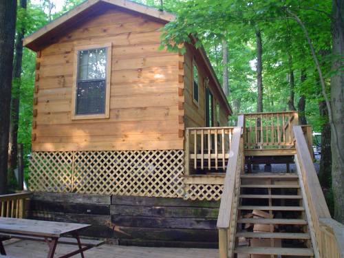 A wooden cabin surrounded by trees, featuring a deck and stairs leading up to the entrance.