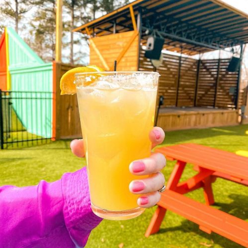A hand holds a refreshing orange drink with a slice of orange, set against a colorful outdoor venue.