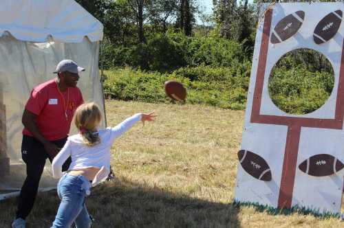 A child throws a football at a target while an adult watches, set in a grassy outdoor area.