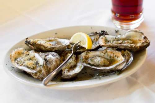 A plate of grilled oysters topped with herbs and a lemon wedge, served with a drink in the background.