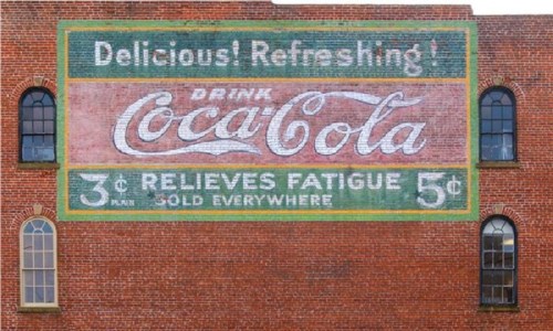 Faded vintage Coca-Cola advertisement on a brick wall, featuring prices and slogans promoting refreshment.