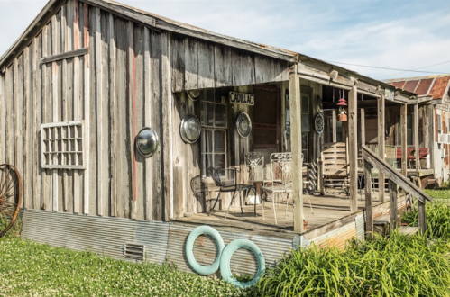 A rustic wooden house with a porch, surrounded by green grass and vintage decor, under a clear blue sky.