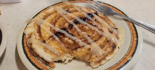 A cinnamon roll pancake drizzled with icing, partially eaten, on a plate with a fork beside it.