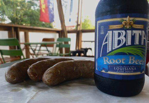A bottle of Abita Root Beer next to three sausages on a table, with outdoor seating in the background.