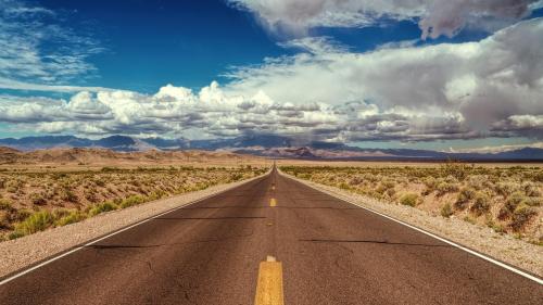 A long, empty road stretches into the distance, surrounded by desert landscape and dramatic clouds above.