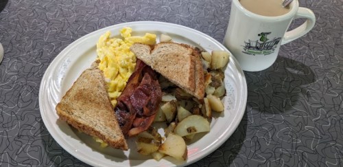 A plate with scrambled eggs, bacon, toast, and potatoes, alongside a cup of coffee on a patterned table.