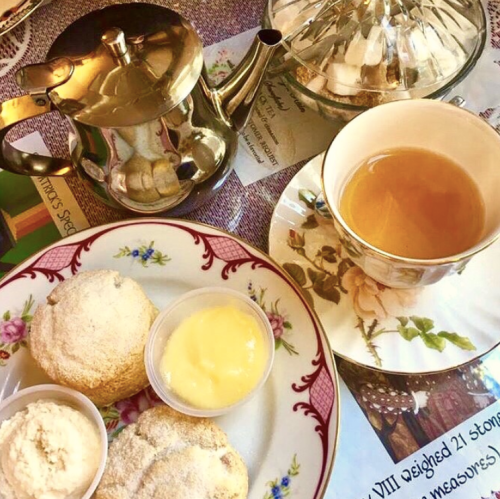A silver teapot, a cup of tea, and two scones with cream and lemon curd on a floral plate.