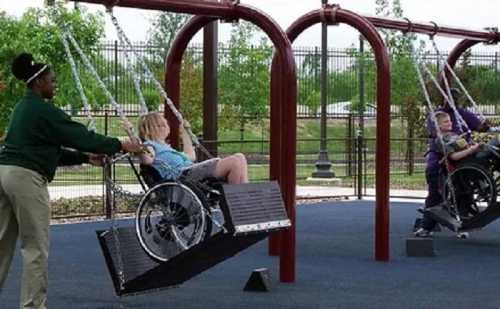 A caregiver pushes a child in a wheelchair on a swing in an accessible playground, with another child in a wheelchair on a swing.