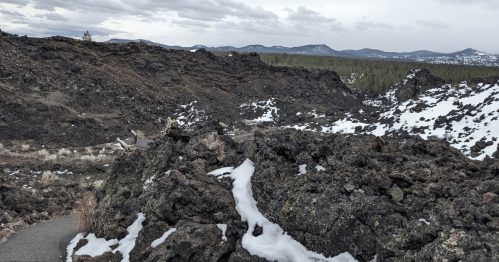 A rugged landscape of dark volcanic rock with patches of snow, surrounded by distant mountains under a cloudy sky.