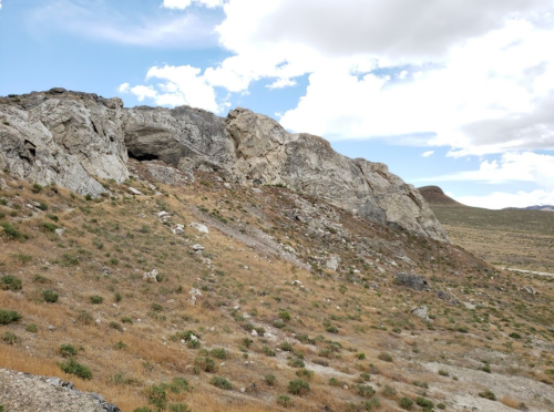 A rocky hillside with sparse vegetation under a partly cloudy sky. A cave entrance is visible in the rock formation.