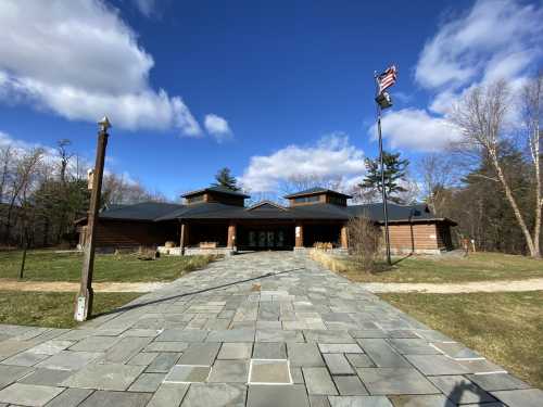 A modern wooden building with a stone pathway, surrounded by grass and trees, under a blue sky with clouds.