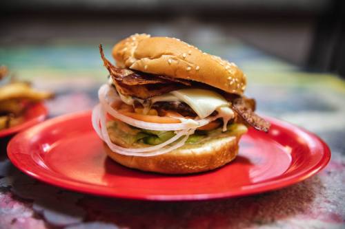 A close-up of a sesame seed bun burger with bacon, cheese, lettuce, tomato, and onion on a red plate.