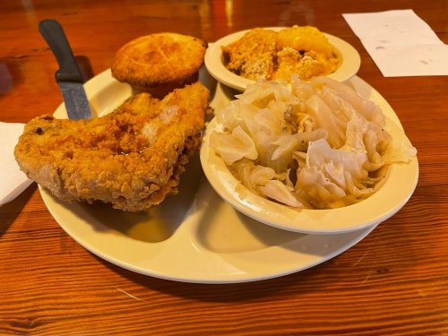 A plate of fried chicken, cornbread, and two sides of cabbage and a creamy dish, served on a wooden table.
