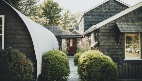 A pathway leads to a red door between two dark-shingled houses, surrounded by lush greenery and trees.