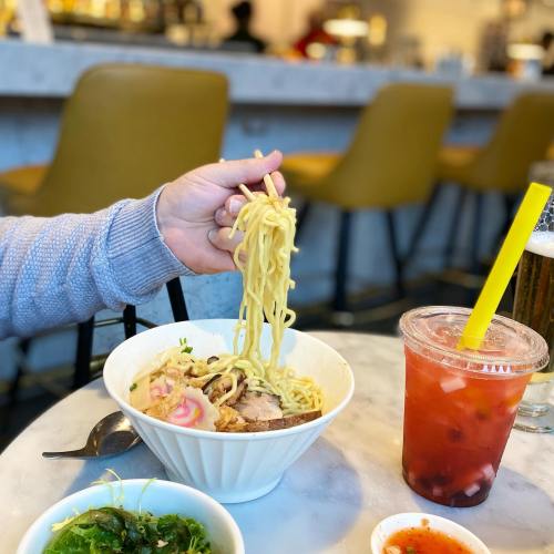 A hand holds a bowl of ramen with noodles, alongside a drink and a small dish of greens on a table.