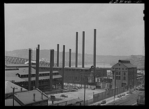 Black and white image of an industrial site with tall smokestacks, buildings, and a river in the background.