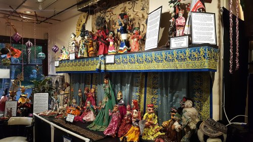 A display of colorful traditional dolls and puppets arranged on a table, with decorative fabrics and informational signs.